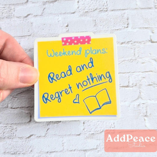 Hand holding a yellow sticker with text 'Weekend plans: Read and Regret nothing' on a white brick wall background.