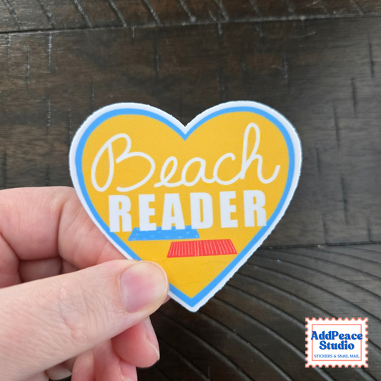 A hand holds a heart-shaped sticker that reads 'Beach READER' in white lettering on a yellow background, with a blue border and a small red-and-blue book graphic. The sticker is photographed against a dark wood surface, and an 'AddPeace Studio' logo appears in the bottom right corner.
