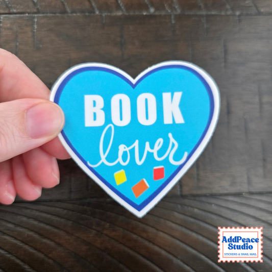 A hand holds a heart-shaped sticker that reads 'BOOK lover' in white text on a bright blue background, with small colored squares near the bottom, set against a dark wood surface. AddPeace Studio logo is visible in the bottom right corner.