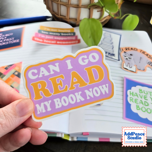 A hand holds a die-cut sticker that reads 'CAN I GO READ MY BOOK NOW' in purple, orange, and white. The sticker is shown in front of an open book with colorful bookmarks, with a plant in the background.