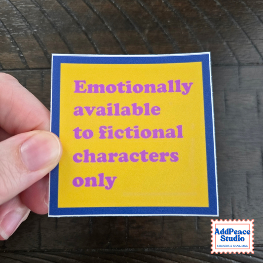 A hand holds a square yellow sticker with a blue border and purple text that reads 'Emotionally available to fictional characters only' against a dark wood background. AddPeace Studio logo is visible in the bottom right corner.