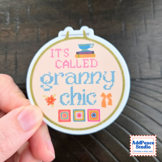 A hand holding a round sticker that reads 'IT'S CALLED granny chic' in pink and blue lettering, with small decorative illustrations, against a dark wood background. AddPeace Studio logo is visible in the bottom right corner.