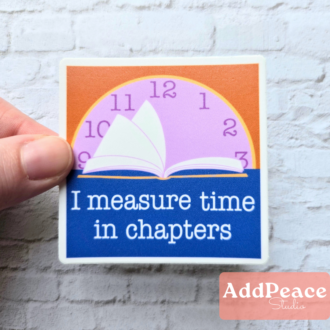 Sticker with a book and clock design, text 'I measure time in chapters', held by a hand on a textured surface.