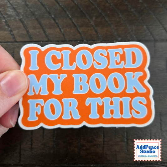 A hand holding a die-cut sticker with the text 'I CLOSED MY BOOK FOR THIS' in light blue bubble letters on an orange background with a white border, set against a dark wood surface. A 'AddPeace Studio' logo is visible in the bottom right corner.