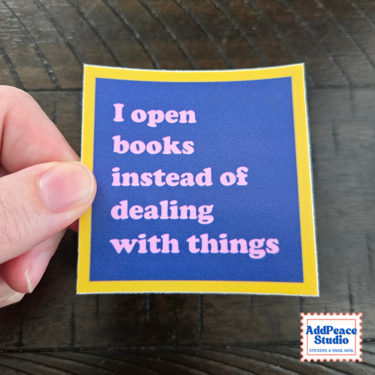 A hand holds a square sticker with a blue background, yellow border, and pink text that reads, 'I open books instead of dealing with things.' The sticker is photographed over a dark wood surface, with an AddPeace Studio logo in the bottom right corner.