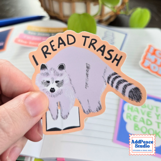 A hand holds a die-cut sticker featuring a raccoon reading a book with the text 'I READ TRASH' in black letters on an orange border. The sticker is shown over an open notebook with other colorful stickers in the background.