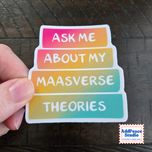 A hand holds a die-cut sticker with a rainbow gradient background and white text reading 'ASK ME ABOUT MY MAASVERSE THEORIES'. A small 'AddPeace Studio' logo appears in the bottom right corner over a dark wood surface.