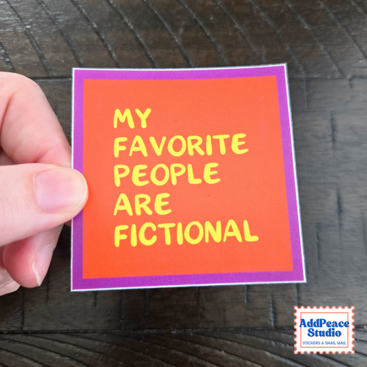 A hand holds a square sticker with a bright orange center, purple border, and yellow text that reads 'MY FAVORITE PEOPLE ARE FICTIONAL' against a dark wood background. An 'AddPeace Studio' logo is visible in the bottom right corner.
