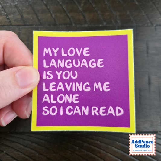 A hand holding a square purple and yellow sticker with white text that reads 'MY LOVE LANGUAGE IS YOU LEAVING ME ALONE SO I CAN READ' on a dark wood background. An 'AddPeace Studio' logo is visible in the bottom right corner.