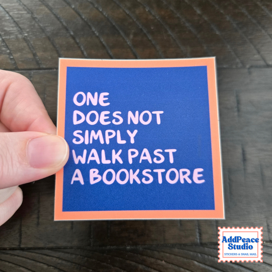 A hand holds a square sticker with a blue background and orange border, featuring the text 'ONE DOES NOT SIMPLY WALK PAST A BOOKSTORE' in white. The sticker is photographed on a dark wood surface, with an 'AddPeace Studio' logo in the bottom right corner.