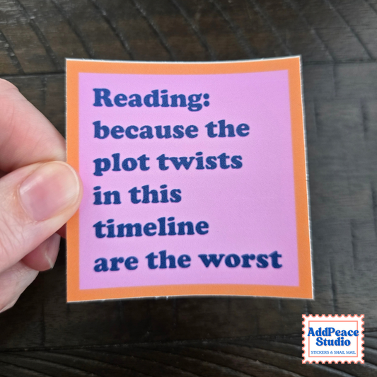 A hand holds a square sticker with a pink background, orange border, and blue text that reads, 'Reading: because the plot twists in this timeline are the worst.' The sticker is photographed over a dark wood surface, with an AddPeaceStudio logo in the bottom right corner.