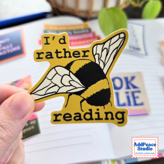 A hand holds a die-cut sticker featuring a yellow and black bumblebee with the text 'I'd rather reading' in black on a yellow background. The background is a blurred desk scene with notebooks and stationery, and an 'AddPeace Studio' logo is visible in the bottom right corner.