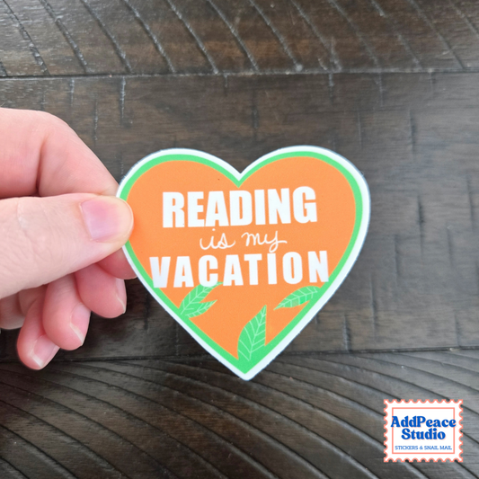 A hand holds a heart-shaped sticker with the text 'READING is my VACATION' in white on an orange background with green leaf accents, set against a dark wood surface. AddPeace Studio logo is visible in the bottom right corner.