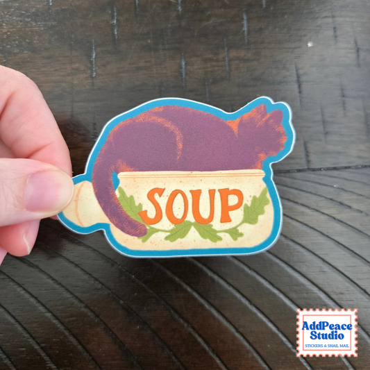 A hand holds a die-cut sticker shaped like a soup bowl, featuring a purple cat sitting inside the bowl with the word 'SOUP' in orange letters and green leaf accents, set against a dark wood background.