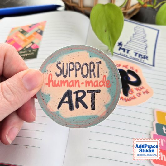 A hand holds a round sticker that reads 'SUPPORT human-made ART' in black lettering over a peach and teal watercolor-style background. The scene is a styled desk setup with an open notebook, other stickers, a plant leaf, and a visible AddPeace Studio logo in the corner.