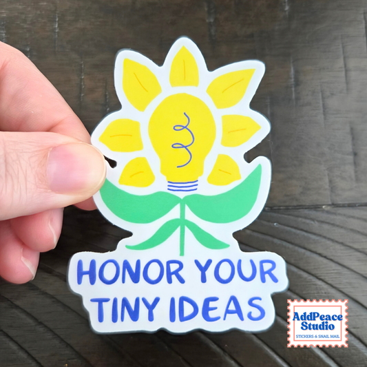 A hand holds a die-cut sticker featuring a yellow sunflower with a lightbulb in the center and green leaves, above the words “HONOR YOUR TINY IDEAS” in blue. The sticker is photographed on a dark wood surface, with an AddPeace Studio logo in the bottom right corner.