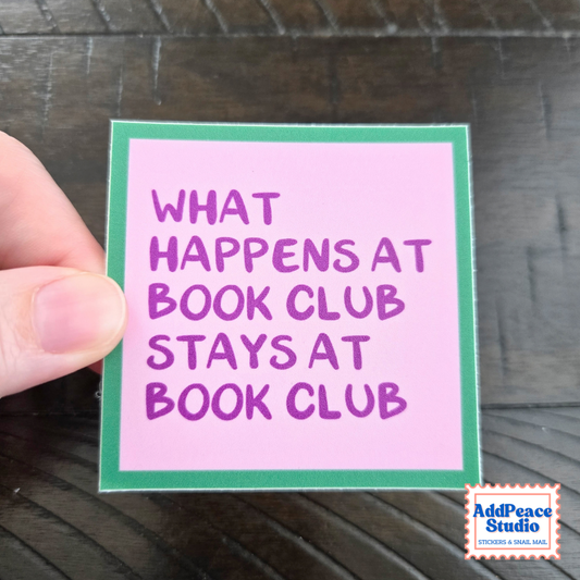 A square pink sticker with a green border held by a hand, featuring the text 'WHAT HAPPENS AT BOOK CLUB STAYS AT BOOK CLUB' in purple. The sticker is photographed on a dark wood surface, with an 'AddPeace Studio' logo in the bottom right corner.