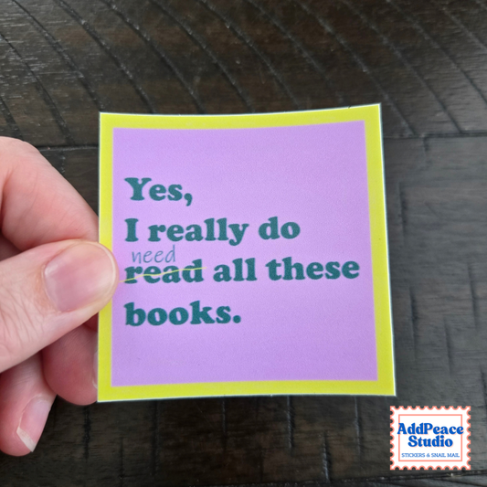 A hand holding a small square sticker with a light purple background, yellow border, and dark teal text that reads, 'Yes, I really do need read all these books.' The sticker is photographed over a dark wood surface, with an AddPeace Studio logo in the bottom right corner.