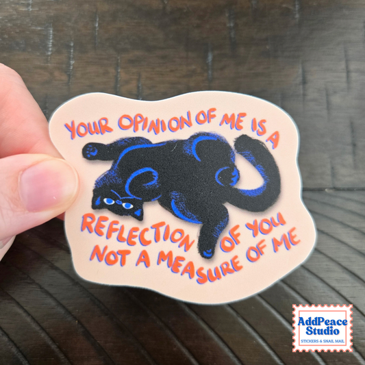 A hand holds a die-cut sticker with a black cat illustration and orange text on a beige background, photographed over a dark wood surface. The sticker reads, "YOUR OPINION OF ME IS A REFLECTION OF YOU NOT A MEASURE OF ME.
