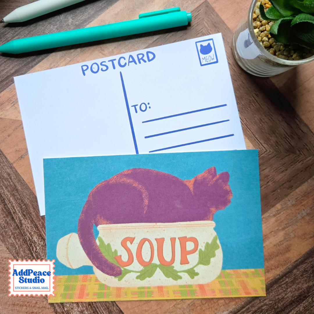 A vintage-style postcard featuring a purple cat curled up inside a soup bowl with the word 'SOUP' on the front, placed on top of a blank postcard on a wooden desk. A teal pen, a small potted succulent, and an AddPeace Studio logo watermark are visible.