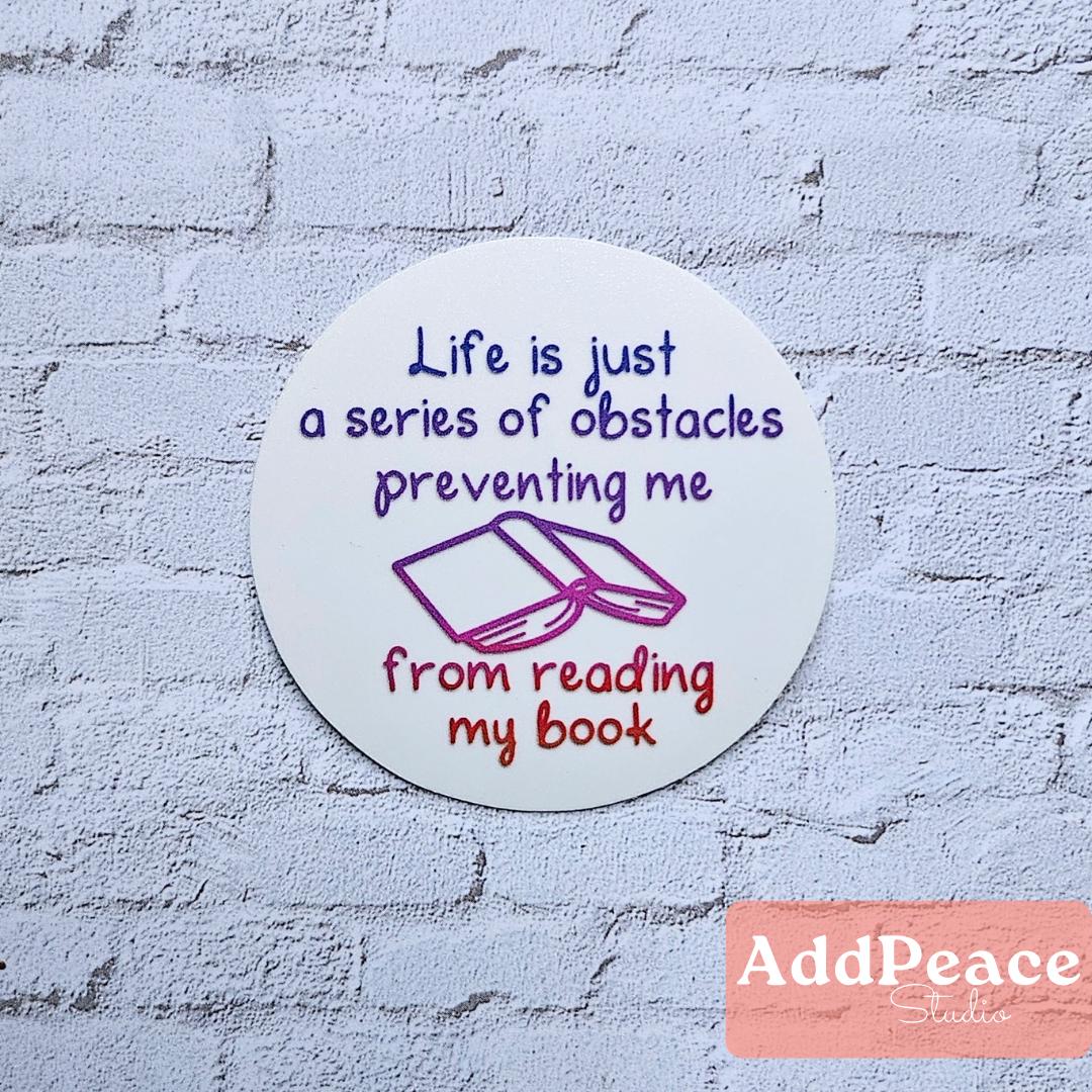 Circular sticker with Life is a series of obstacles preventing me from reading my book on a textured wall, featuring the brand 'AddPeace'