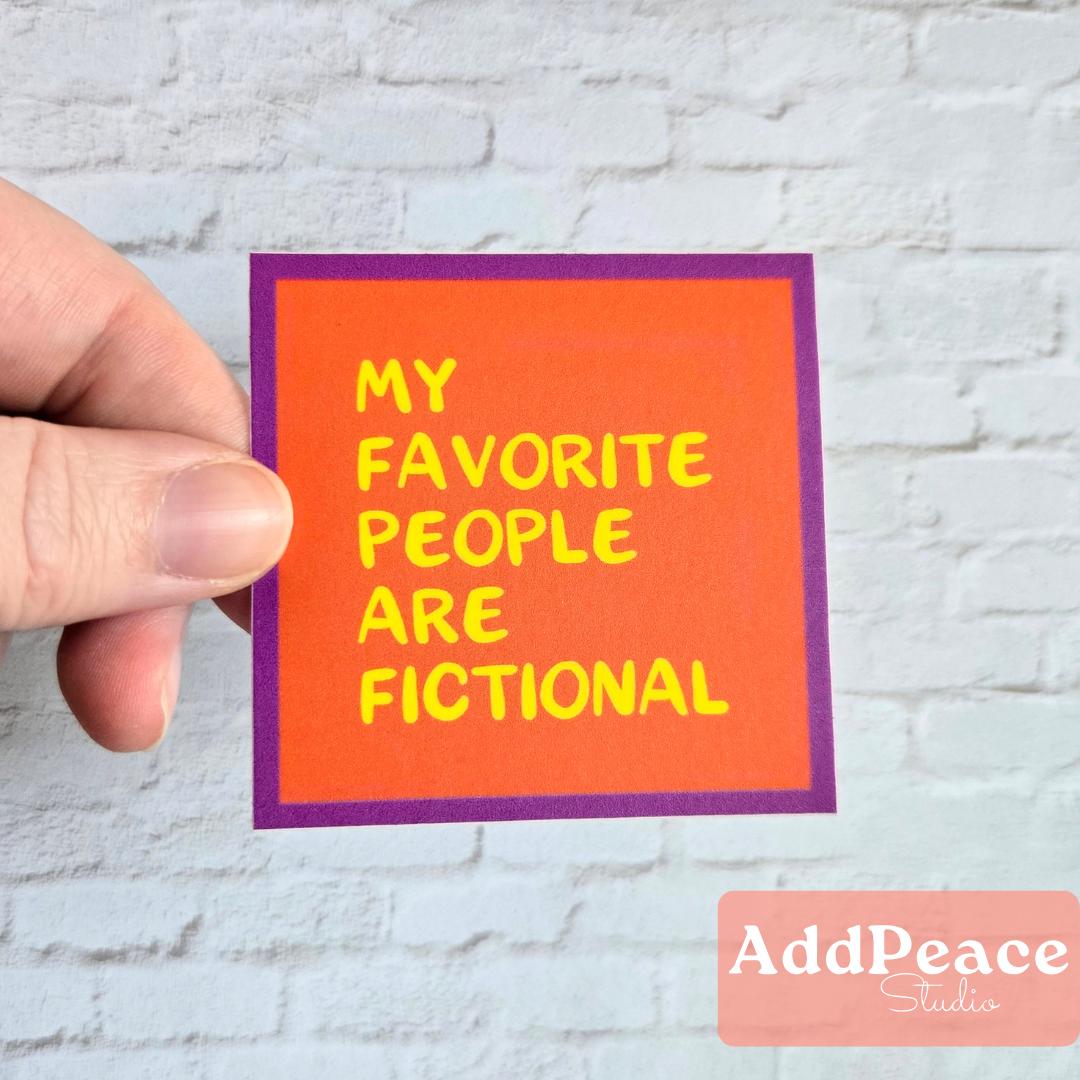 Hand holding a colorful sticker with 'My favorite people are fictional' text against a light brick wall.