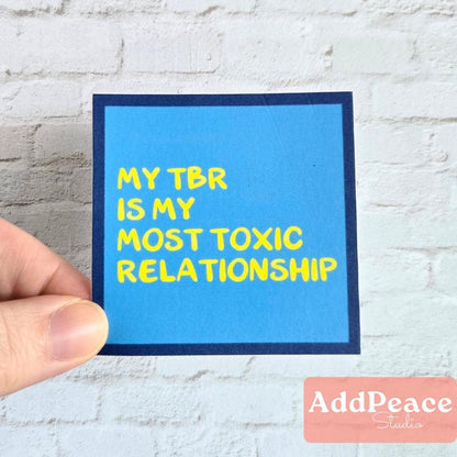 Blue sticker with yellow text 'MY TBR IS MY MOST TOXIC RELATIONSHIP' held by a hand against a white brick wall.