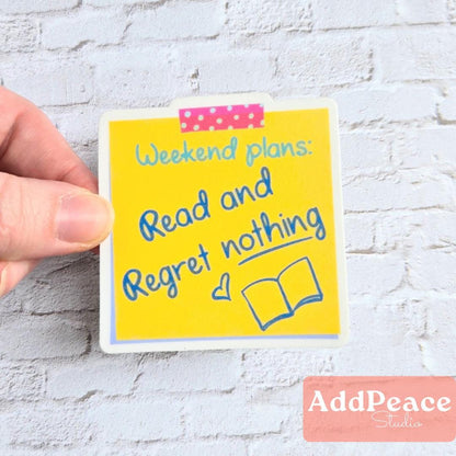Hand holding a yellow sticker with text 'Weekend plans: Read and Regret nothing' on a white brick wall background.