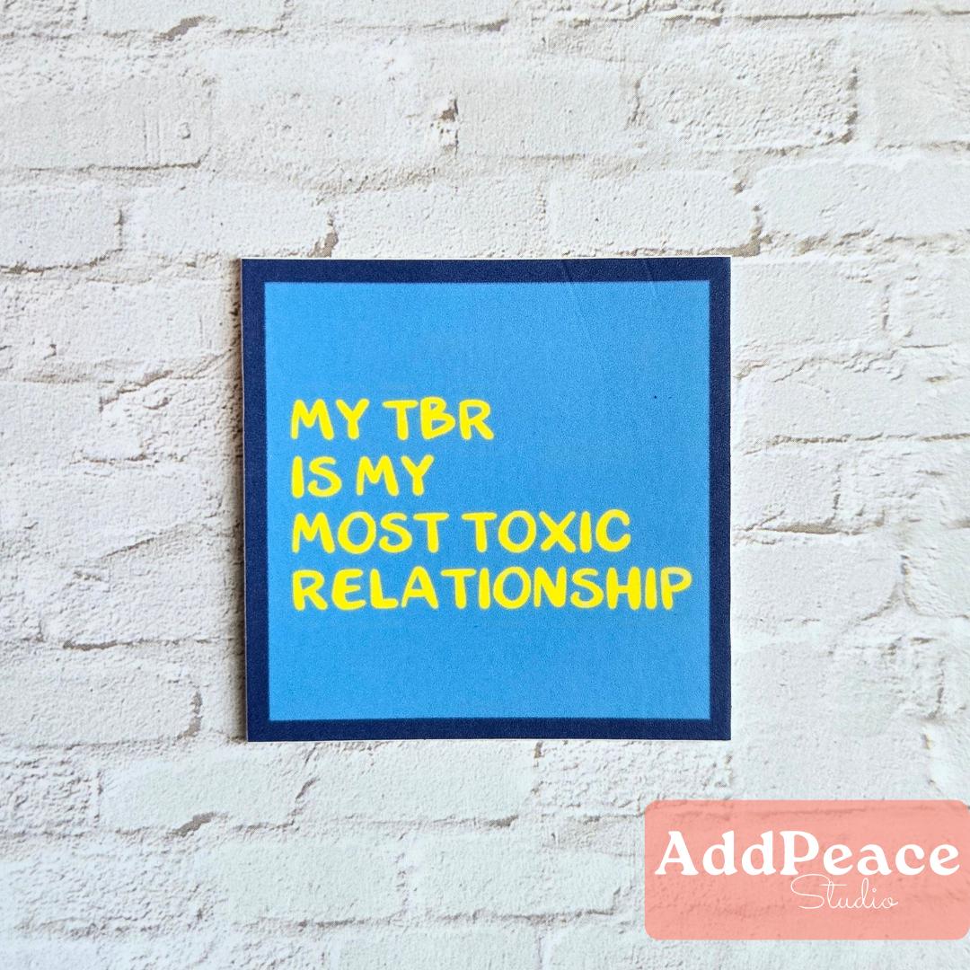 Blue sticker with text 'My TBR is my most toxic relationship' on a white brick wall, with 'AddPeace Studios' logo at the bottom.