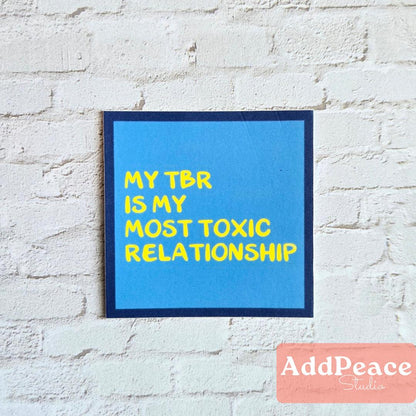 Blue sticker with text 'My TBR is my most toxic relationship' on a white brick wall, with 'AddPeace Studios' logo at the bottom.