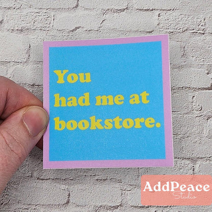 Hand holding a blue sticker with yellow text You had me at bookstore, against a textured wall, featuring the brand 'AddPeace'.