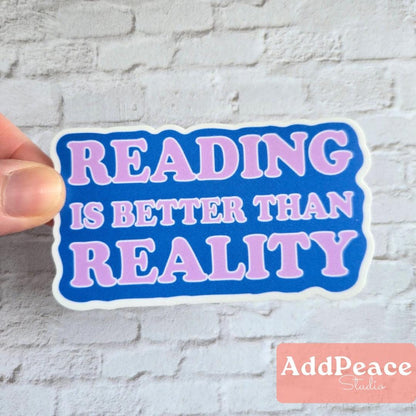 Sticker with 'READING IS BETTER THAN REALITY' text held against a white brick wall, with 'AddPeace Studios' branding.