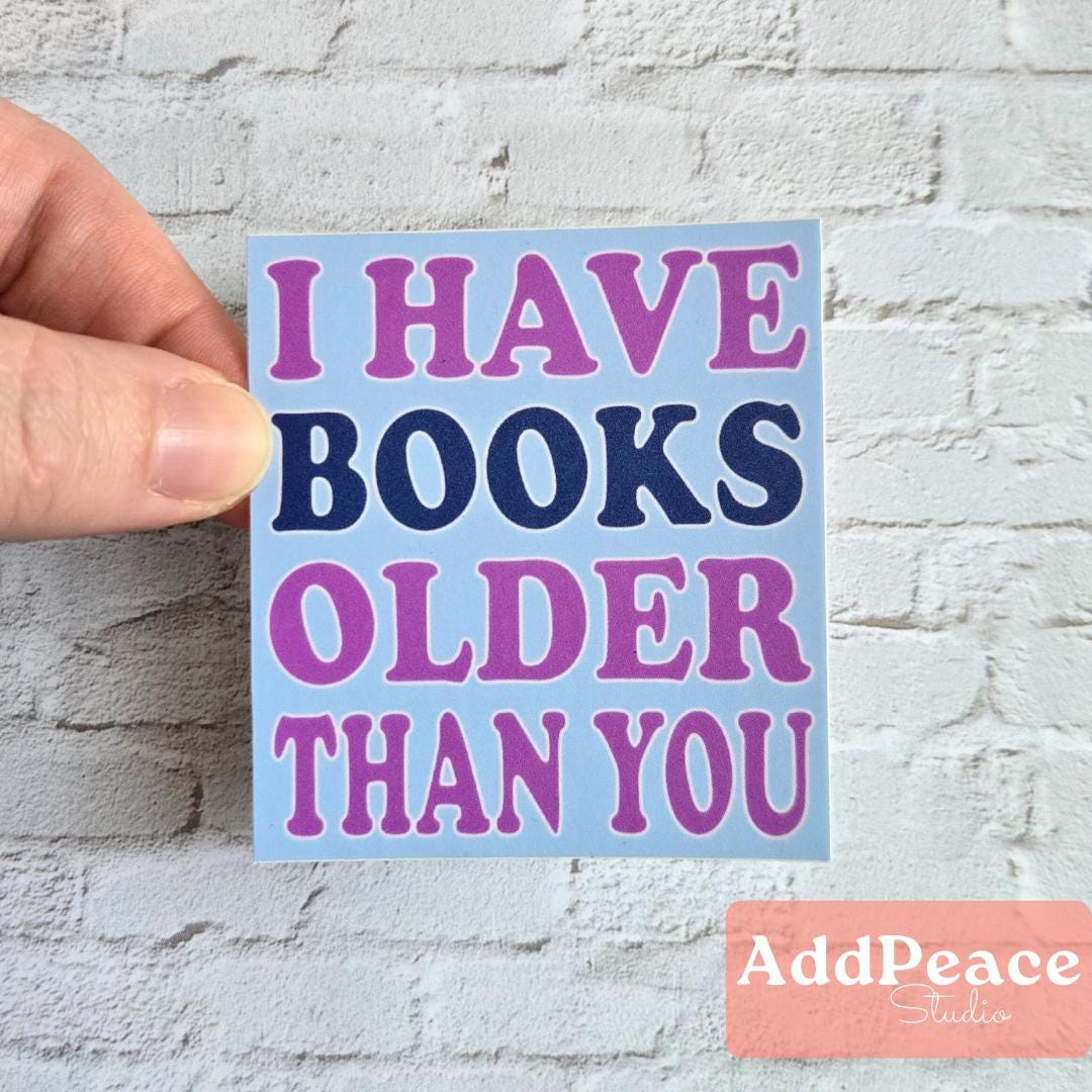 Hand holding a sticker with text 'I HAVE BOOKS OLDER THAN YOU' on a light blue background.
