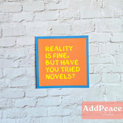 Orange sticker with Reality is fine but have you tried novels? on a brick wall with 'AddPeace Studios' branding.