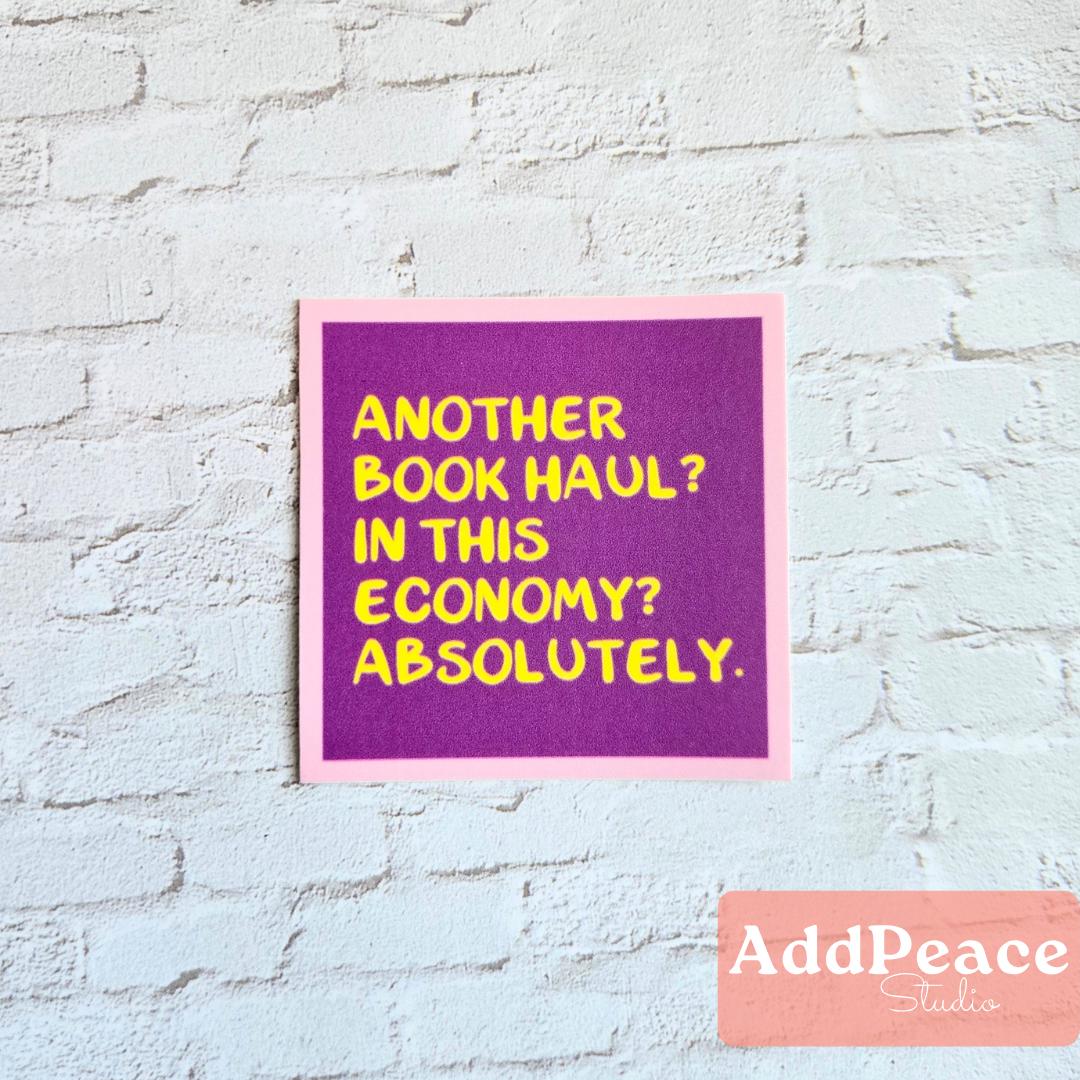 Purple sticker with yellow text Another book haul in this economy? Absolutely. on a light blue brick wall, featuring the brand 'AddPeace Studios'.