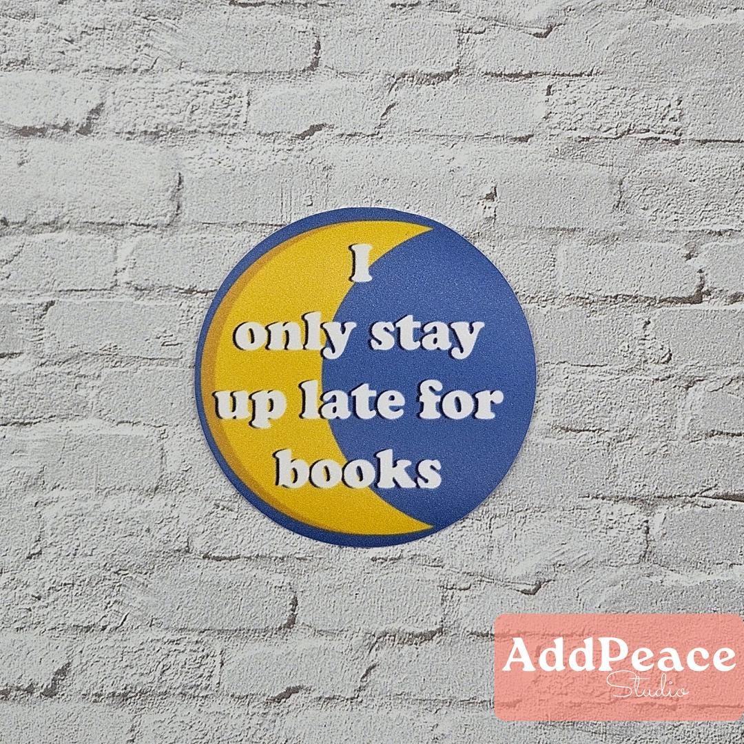 Circular sticker with 'I only stay up late for books' text on a yellow and blue design against a gray brick wall, with 'AddPeace' branding at the bottom.