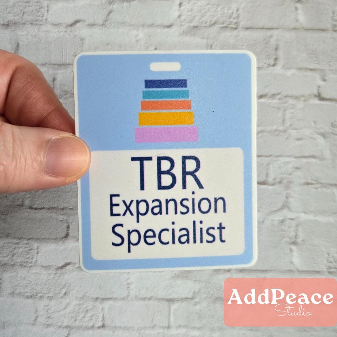 Hand holding a sticker labeled 'TBR Expansion Specialist' with colorful design on a gray brick wall background.