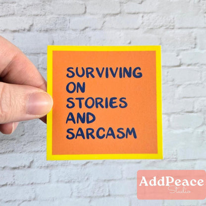 Hand holding an orange sticker with text 'Surviving on Stories and Sarcasm' against a light brick wall.