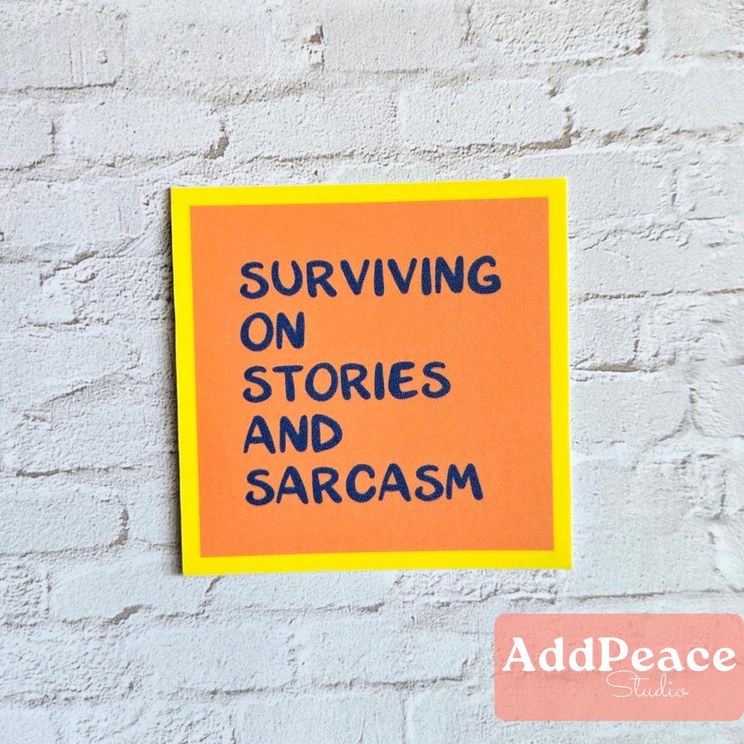 Orange sticker with blue text 'Surviving on Stories and Sarcasm' on a white brick wall, featuring AddPeace Studios logo.