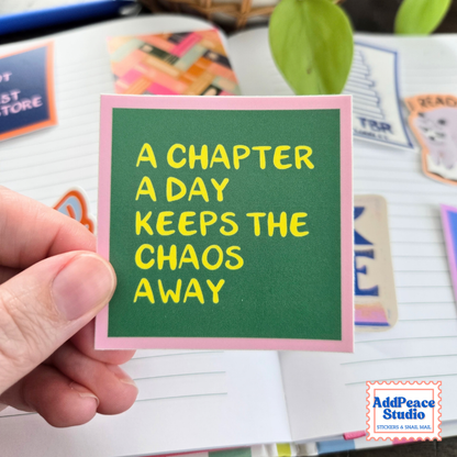 A hand holds a square sticker with a green chalkboard-style center, pink border, and yellow text reading 'A CHAPTER A DAY KEEPS THE CHAOS AWAY'. The background shows a notebook and other colorful stickers, with an AddPeace Studio logo in the bottom right corner.