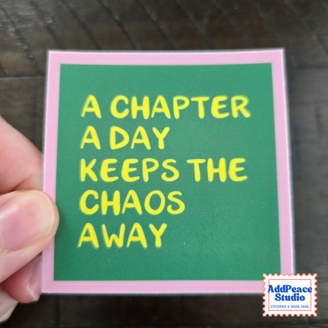 A hand holding a square sticker with a green background, pink border, and yellow text that reads 'A CHAPTER A DAY KEEPS THE CHAOS AWAY'. A small 'AddPeace Studio' logo is in the bottom right corner.