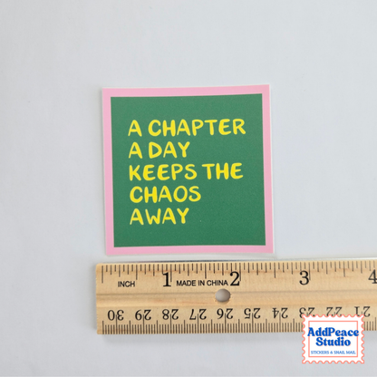 A square sticker with the text 'A CHAPTER A DAY KEEPS THE CHAOS AWAY' in yellow on a green background with a pink border, shown above a wooden ruler for scale on a light gray background.