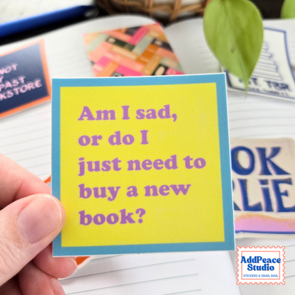 A hand holds a square yellow sticker with a blue border and purple text that reads, 'Am I sad, or do I just need to buy a new book?'. The background shows a notebook, other colorful stickrtd and a green leaf.