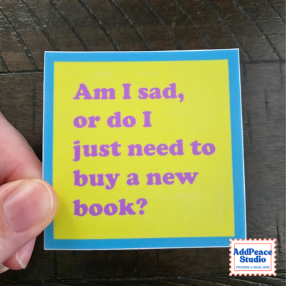 A hand holding a square yellow sticker with a blue border and purple text that reads, 'Am I sad, or do I just need to buy a new book?' on a dark wood background. An 'AddPeace Studio' logo is visible in the bottom right corner.