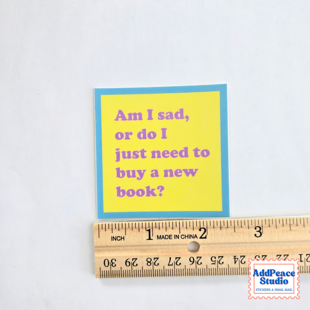 A small square sticker with a yellow center and blue border, reading 'Am I sad, or do I just need to buy a new book?' in purple text, shown next to a wooden ruler on a white background. A small 'AddPeace Studio' watermark appears in the lower right corner.