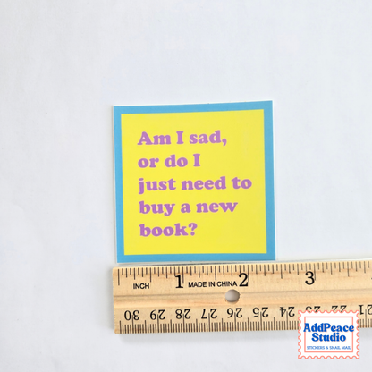 A small square sticker with a yellow center and blue border, reading 'Am I sad, or do I just need to buy a new book?' in purple text, shown next to a wooden ruler on a white background. A small 'AddPeace Studio' watermark appears in the lower right corner.