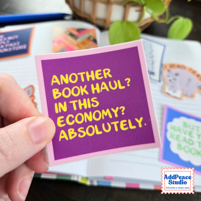 A hand holds a square sticker with purple background and light pink border with yellow text that says Another book haul? In this economy? Absolutely. The background has an open notebook with colorful stickers and plants.