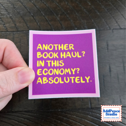 A hand holds a square purple sticker with yellow text that reads 'ANOTHER BOOK HAUL? IN THIS ECONOMY? ABSOLUTELY.' against a dark wood background.