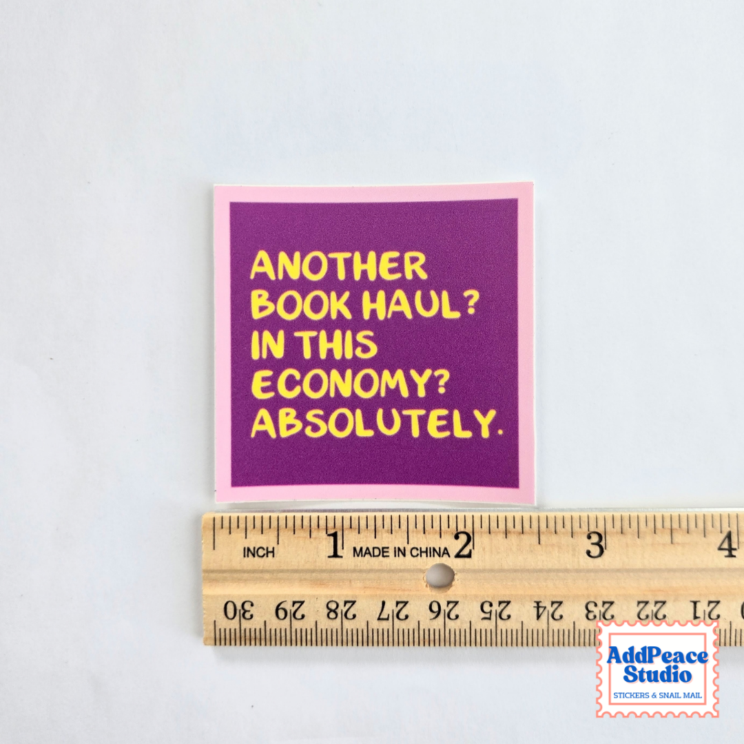 A square sticker with a purple background and yellow text reading 'ANOTHER BOOK HAUL? IN THIS ECONOMY? ABSOLUTELY.' with a pink border, shown above a wooden ruler for scale on a white background.