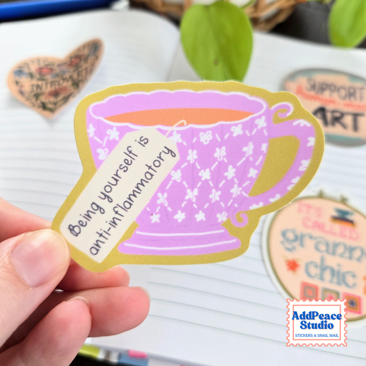 A hand holds a die-cut sticker shaped like a lavender teacup with white floral patterns, a gold outline, and a small tag reading 'Being yourself is anti-inflammatory.' The background shows a notebook, decorative stones, and a blurred green plant, with an 'AddPeace Studio' logo in the bottom right corner.